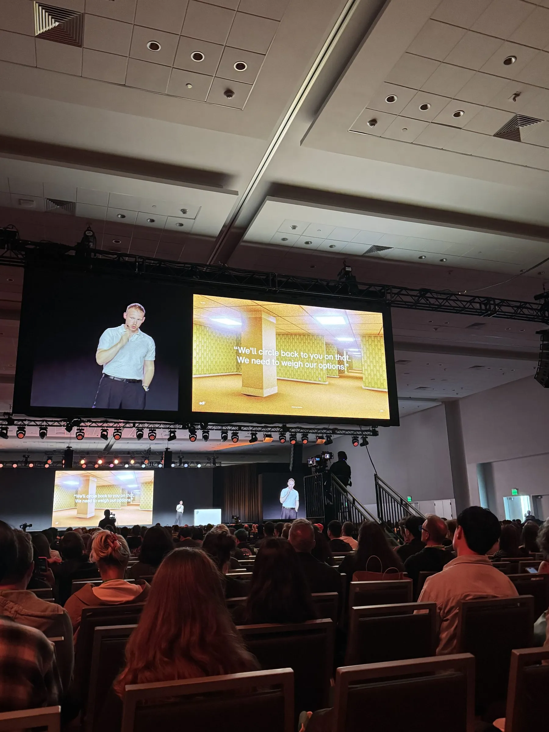 A speaker on the main stage, the screen beside him showing a beige office corridor captioned "We'll circle back to you on that. We need to weigh our options."
